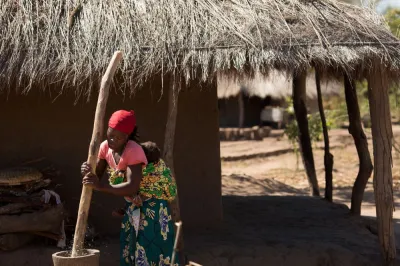 an African woman with a baby on her back works a large mortar